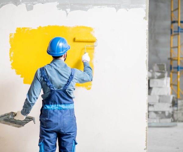 Workman in uniform painting wall with yellow paint at the construction site indoors